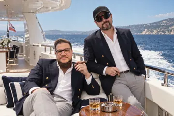 Aaron Rountree and Andrew Lee Jenkins in navy blazers with cigars on the deck of a yacht at sea