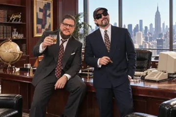 Aaron Rountree raising a glass and Andrew Lee Jenkins holding a cigar in a corner office with a city skyline and a beige 1990s computer